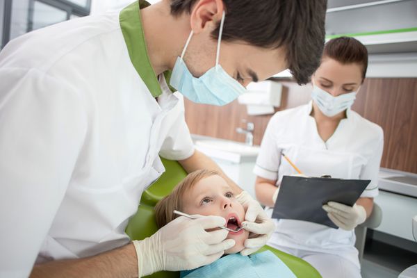 دندانپزشک جوان شاد در حال معاینه دندان های بیمار کوچک است او ابزارها را در دست گرفته و با جدیت به کودک نگاه می کند پسر با تلاش دهانش را باز می کند دستیار زن در حال نوشتن یادداشت است