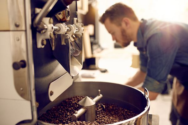 دانه‌های قهوه تیره و معطر در دستگاه روست مدرن با تصویر تار قهوه‌پز حرفه‌ای در پس‌زمینه قابل مشاهده است