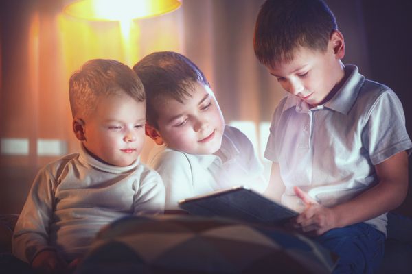 سه کودک در شب در اتاق خواب از رایانه لوحی استفاده می کنند برادران با رایانه لوحی در یک اتاق تاریک بچه های پسر بچه در حال تماشای فیلم با هم زمان خواب