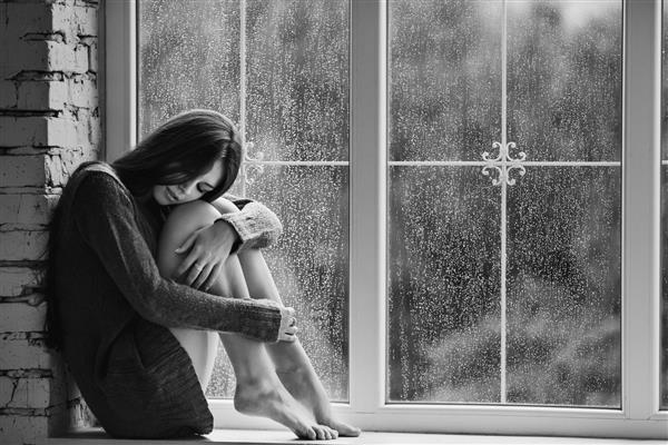 زن جوان زیبا تنها با قطرات باران نزدیک پنجره نشسته است و دختر غمگین مفهوم تنهایی تصویر سیاه و سفید