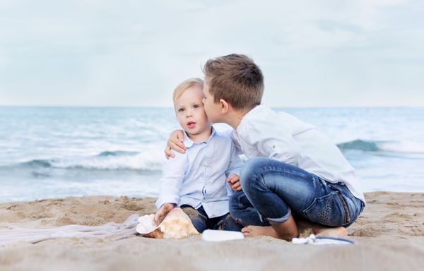 برادر بزرگتر خوشحال برادر کوچک و ناز خود را در ساحل می بوسد کودکانی که در ساحل با ماسه بازی می کنند مفهوم برادری دوستی