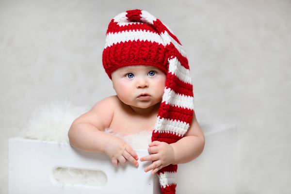 یک نوزاد کریسمس که در جعبه چوبی سفید نشسته است و کلاه دم بلند راه راه قرمز و سفید بر سر دارد