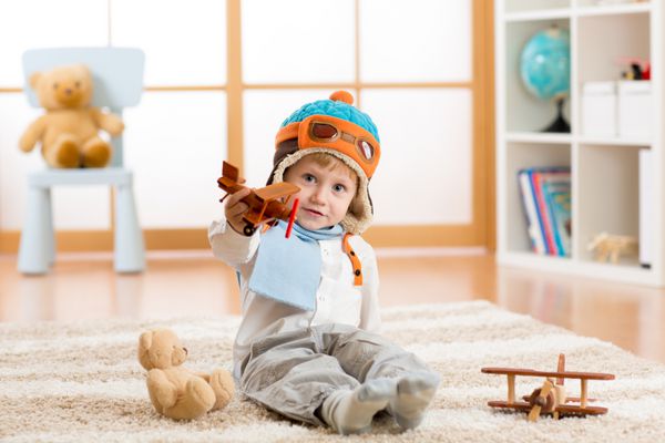 پسر بچه شاد در حال بازی با هواپیمای چوبی اسباب بازی و خرس عروسکی روی زمین در اتاق مهد کودک