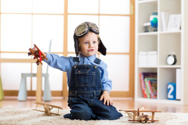 کودکی که مانند خلبان خلبان لباس می پوشد در خانه در اتاقش با هواپیماهای اسباب بازی بازی می کند