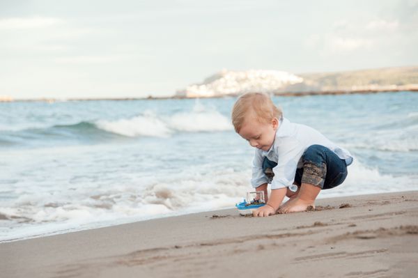 پسر کوچک در حال بازی در ساحل با یک کشتی مفهوم تعطیلات تابستانی و سفر کودکی مبارک
