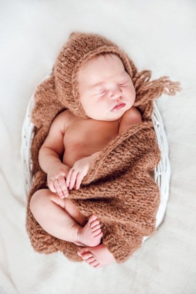 یک نوزاد تازه متولد شده در یک سبد حصیری سفید پوشیده شده با یک پتو بافتنی باقالی قهوه ای خوابیده است