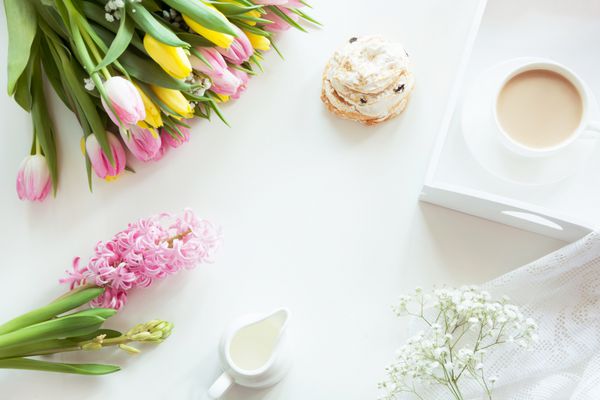 صبحانه صبح در بهار با یک فنجان قهوه سیاه با شیر و شیرینی به رنگ های پاستلی یک دسته گل لاله تازه زرد و صورتی در زمینه سفید نمای بالا