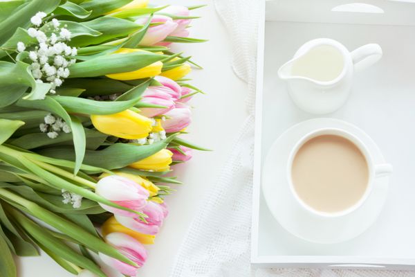 صبحانه صبح در بهار با یک فنجان قهوه سیاه با شیر در رنگ های پاستلی دسته گلی از لاله های تازه زرد و صورتی در زمینه سفید نمای بالا