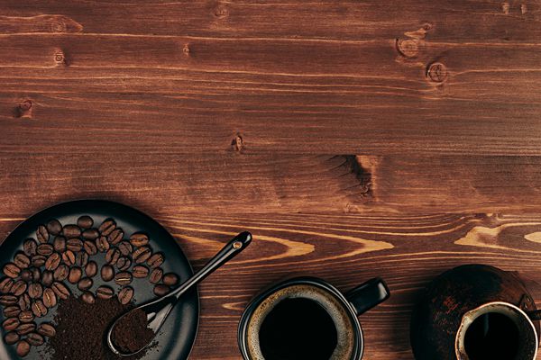 قهوه داغ در فنجان سیاه با دانه‌ها قاشق و دیگ ترکی با فضای کپی روی پس‌زمینه تخته چوبی قدیمی قهوه‌ای نمای بالا سبک روستیک