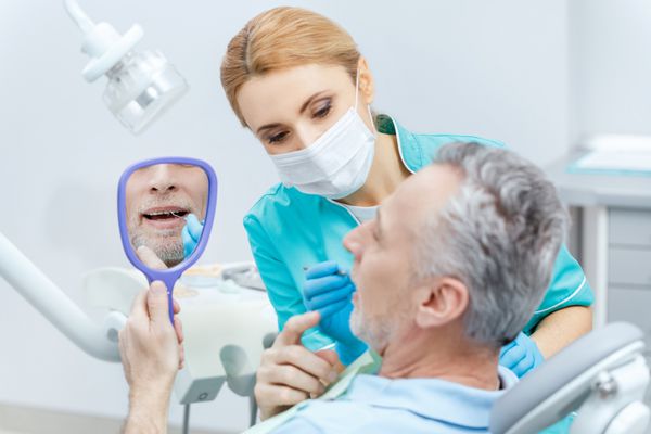 بیمار بالغ در حالی که دندانپزشک حرفه ای در حال بررسی دندان ها است به آینه نگاه می کند