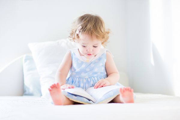 دختر نوپا بامزه با لباس آبی در حال خواندن کتاب روی تخت سفید