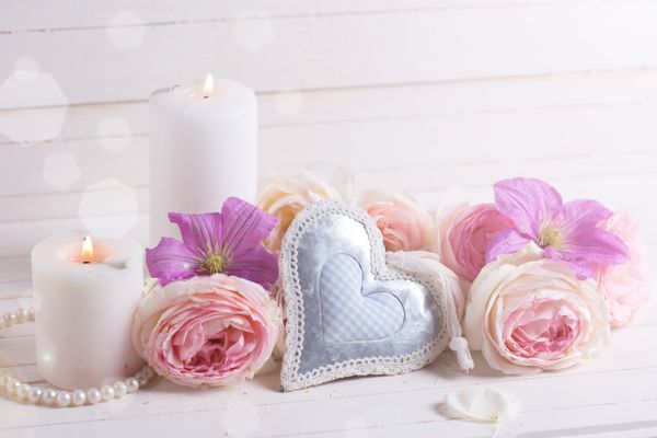 قلب تزئینی گل رز صورتی و گل های کلماتیس بنفش شمع در پرتو نور در زمینه چوبی سفید تمرکز انتخابی مکانی برای متن