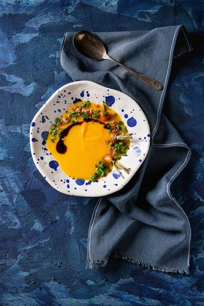 سوپ سبزیجات کدو و هویج سرو شده با قاشق و سس بالزامیک و پیاز سرخ شده روی دستمال آبی تیره
