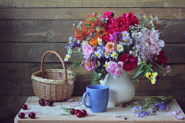 طبیعت بی جان با یک دسته گل تابستانی با گل صد تومانی رز میخک و گل های دیگر در یک کوزه سفید و گیلاس های رسیده روی میز چوبی