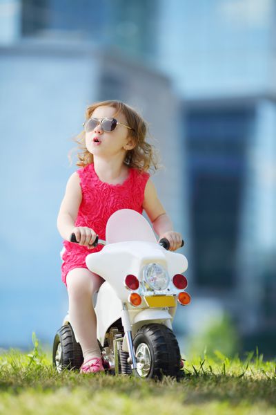 دختر کوچک شاد با عینک آفتابی سوار بر موتور اسباب بازی روی چمن نزدیک آسمان خراش ها می شود