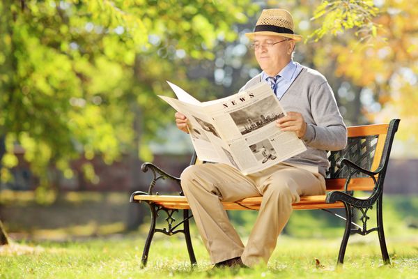 آقای ارشد با کلاه روی یک نیمکت چوبی نشسته و در پارک مشغول خواندن روزنامه است