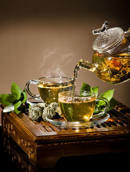 عکس عمودی از قوری شیشه ای جریان چای سبز در فنجان در زمینه قهوه ای مراسم چای