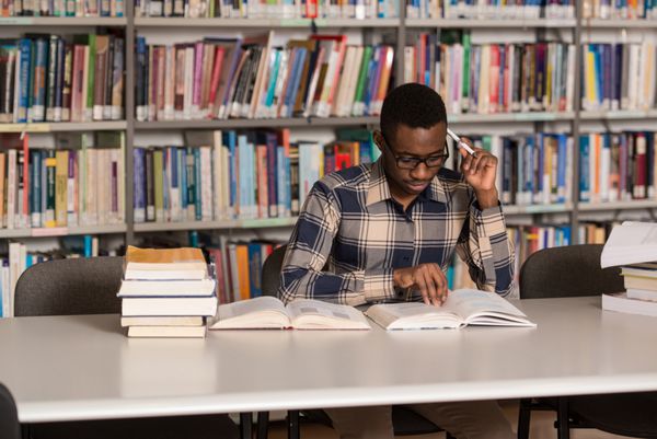 پرتره دانشجوی باهوش آفریقایی با کتاب باز در حال خواندن آن در کتابخانه کالج - عمق میدان کم