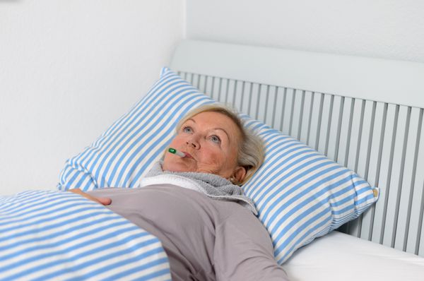 زن میانسال بیمار دراز کشیده روی تخت با دماسنج در دهان در حالی که با حالتی متفکر به بالا نگاه می کند