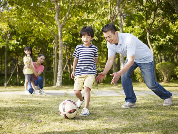 پدر آسیایی در پارکی به پسرش فوتبال فوتبال را آموزش می‌دهد در حالی که مادر و دختر از پشت تماشا می‌کنند