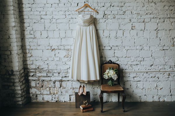 لباس عروس سفید کفش و یک دسته گل سفید و سبز در مقابل یک دیوار آجری سفید در اتاق زیر شیروانی