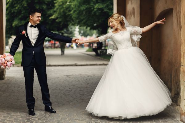 داماد خوش تیپ و عروس زیبا با لباس سفید در حال رقصیدن زیر قوس