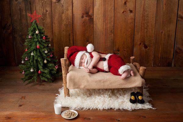 نوزاد دو هفته ای نوزاد پسری که کت و شلوار بابانوئل قلاب بافی پوشیده و روی تخت چوبی کوچکی می خوابد در استودیو با وسایلی از جمله درخت کریسمس لیوان شیر و کلوچه های قلاب بافی گرفته شده است