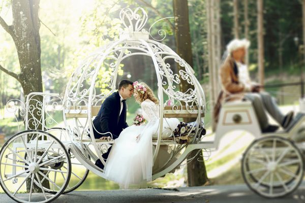 داماد خوش تیپ در حال بوسیدن عروس زیبا در کالسکه افسانه ای جادویی در پارک نور خورشید