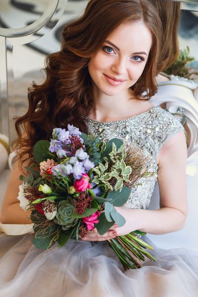 خانم جوان زیبا با لباسی لوکس در فضای داخلی زیبا با دسته گلی که نزدیک آینه نشسته است