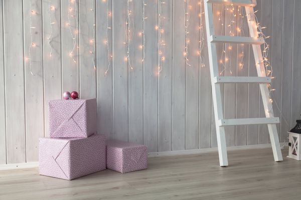 چراغ‌های کریسمس در پس‌زمینه چوبی سفید با جعبه‌های هدیه و پله‌های صورتی می‌سوزند