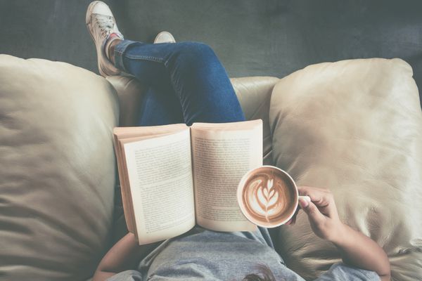 عکس ملایم دختر جوان در حال خواندن کتاب و نوشیدن قهوه نمای بالا