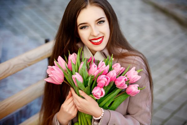 زن جوان زیبا با دسته گل لاله های بهاری در خیابان شهر دختر شادی که لبخند می زند و گل های لاله صورتی را در فضای باز نگه می دارد پرتره بهاری زن زیبا در پارک