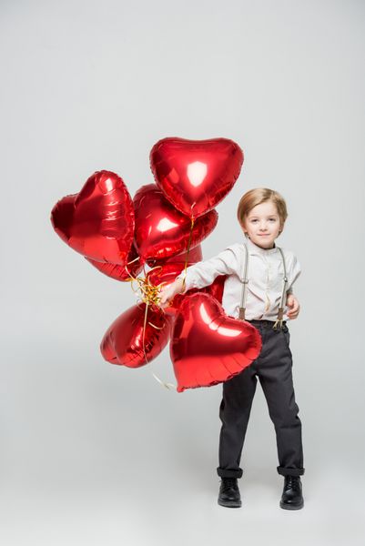 پسر کوچکی که دسته ای از بادکنک های هوایی به شکل قلب قرمز جدا شده روی خاکستری را در دست دارد