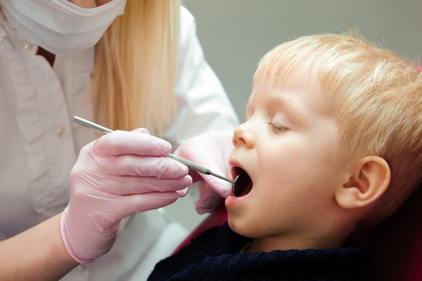 دندانپزشک زن دندان های کودک بیمار را معاینه می کند دهان کودک روی صندلی دندانپزشک کاملا باز است