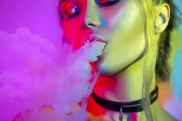پرتره هنری مد از زن مدل زیبایی در نورهای روشن با دود رنگارنگ دختر سیگاری از نزدیک یک زن در حال استنشاق از سیگار الکترونیکی مفهوم زندگی در شب