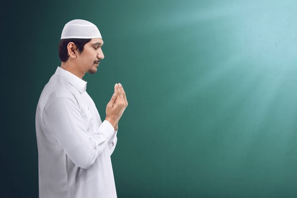 مرد جوان مسلمان آسیایی در حال دعا با خدا لباس مسلمان و کلاه بر سر
