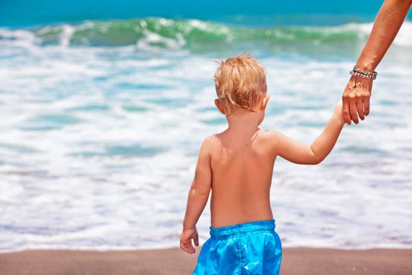 سفر خانوادگی شاد - مادر و پسر بچه با تفریح در ساحل ماسه سیاه قدم می زنند تا برای شنا در دریا موج سواری کنند شیوه زندگی والدین فعال فعالیت افراد در فضای باز در جزیره گرمسیری تعطیلات تابستانی با کودک
