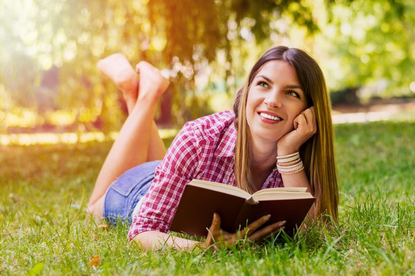 زن جوان و خوش‌نظر روی چمن‌های سبز دراز کشیده و در مقابل پارک شهر کتاب می‌خواند