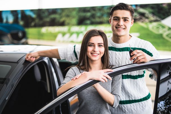 زوج جوان زیبا در حالی که در یک نمایشگاه موتوری به ماشین جدیدشان تکیه داده اند لبخند می زنند و به دوربین نگاه می کنند مردی کلید ماشین را در دست دارد