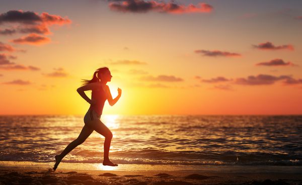 زن دونده در حال دویدن در ساحل هنگام غروب آفتاب