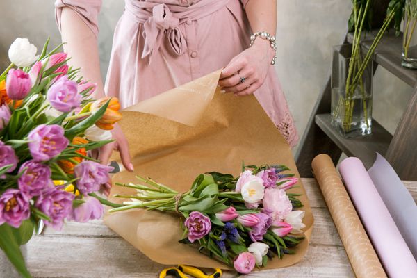 بسته دکوراتور دسته گل جشن در کاغذ بسته بندی گل‌فروش جوان زیبا گل‌فروشی را با گل صد تومانی صورتی و گل‌های وحشی در کارگاه روی زمینه چوبی مونتاژ می‌کند دست زن در محل کار