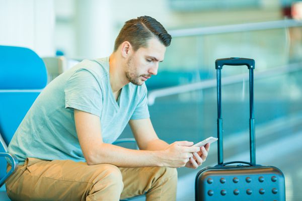 مرد جوانی در سالن فرودگاه منتظر هواپیمای پرواز است مرد قفقازی با گوشی هوشمند در اتاق انتظار