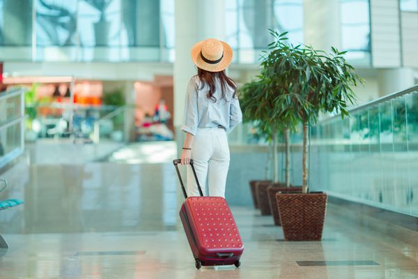 زن جوان با کلاه با چمدان در فرودگاه بین المللی مسافر خطوط هوایی در سالن فرودگاه در انتظار هواپیمای پرواز