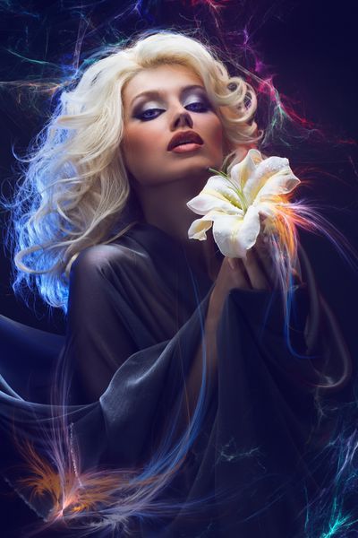 بلوند جذاب با چشمان آبی که زنبق سفید را در دست در پس زمینه نورپردازی انتزاعی نگه می دارد