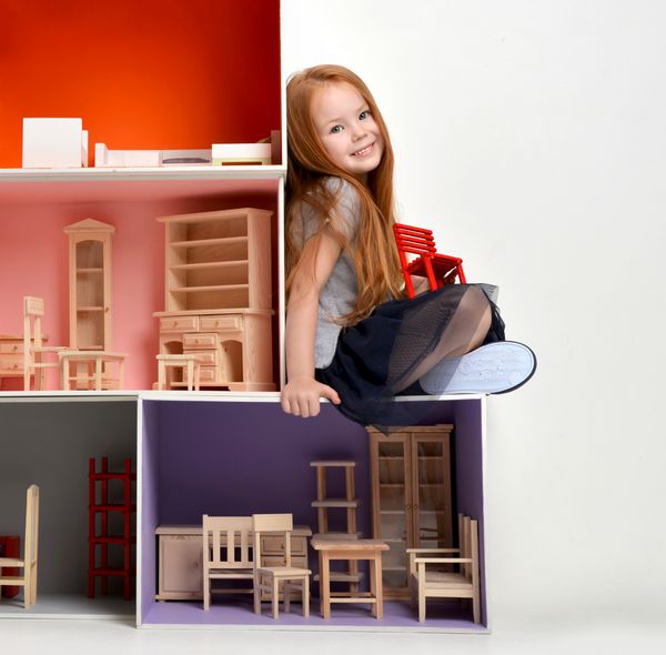 بچه دختر بچه مو قرمز در حال بازی با خانه عروسک پر شده با اسباب بازی های کوچک مبلمان