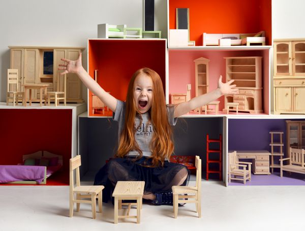 بچه دختر بچه مو قرمز در حال بازی با خانه عروسک پر شده با اسباب بازی های مبلمان کوچک و عروسک