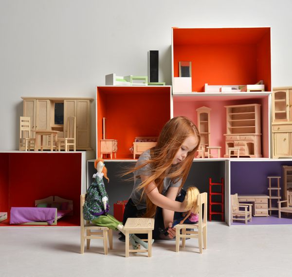 بچه دختر بچه مو قرمز در حال بازی با خانه عروسک پر شده با اسباب بازی های کوچک مبلمان و عروسک نشسته