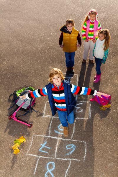 پسر پریدن در بازی hopscotch با دوستان پسرها دختران ایستاده با کیسه های مدرسه در نزدیکی قرار دارد