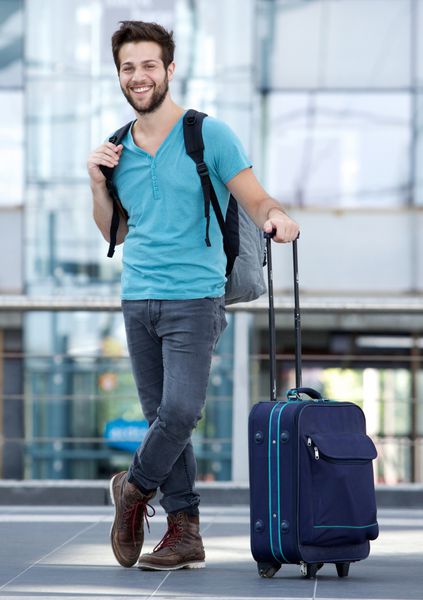 پرتره کامل از یک مرد جوان در فرودگاه با چمدان و کیسه انتظار