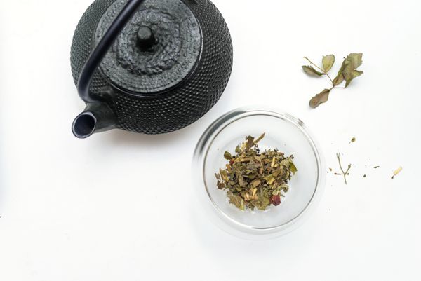 قوری قهوه ای آسیا و فنجان شیشه ای با چای گیاهی گیاهان خشک برگ ها و گل ها نمای بالا جدا شده بر روی زمینه سفید